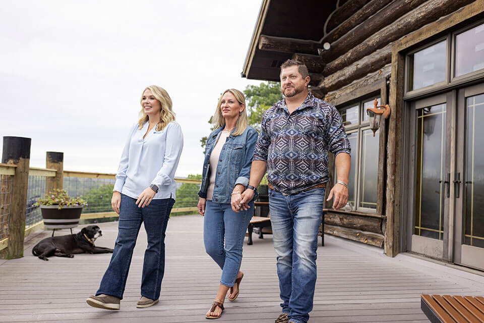 Three people walk across an expansive deck attached to a log home in the country.