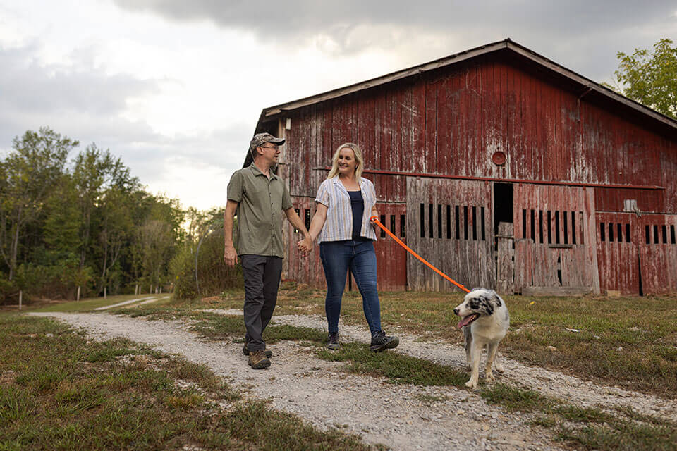 A man and woman walk a dog on a leash past an old barn.