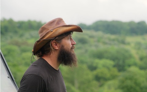 A man, in profile, with a big beard looking off into the distance, with woods beyond him.